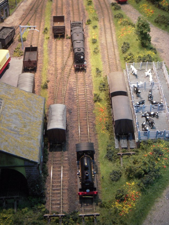 Set in Devon between 1948 and early 1960s and separated from a fictional station - really a fiddle yard - by a bridge, 8' x 2' Yard Shunter saw train engines being released by the eponymous fixed wheelbase locomotives - ranging from a sound fitted BR 08 to a Beattie Well Tank - and the wagons destined for a private timber merchant, goods shed, coal merchants and cattle pen being re-arranged.  Points on the C&L hand built wooden sleeper track were worked by under-board servo point operation controlled ultimately via MERG servo 4 and accessory decoder.  Meanwhile NCE PowerCab DCC also controlled ex Southern Railway N and Q1 Class locomotives as well as a Standard 4MT 2-6-4T.